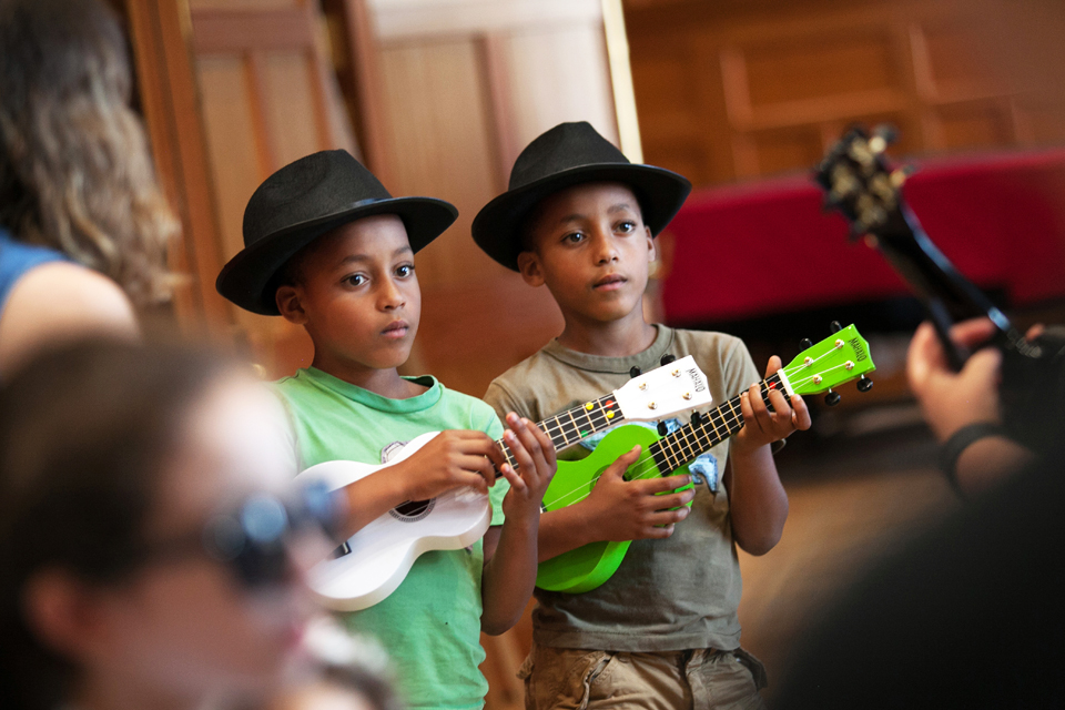 Two black boys, wearing black hats, wearing green shirts and playing the ukulele.